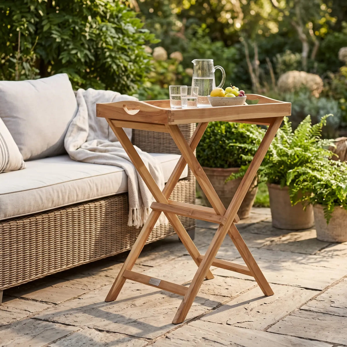 Teak Tray Table with folding frame beside a wicker sofa on a terrace with a water pitcher and fruit